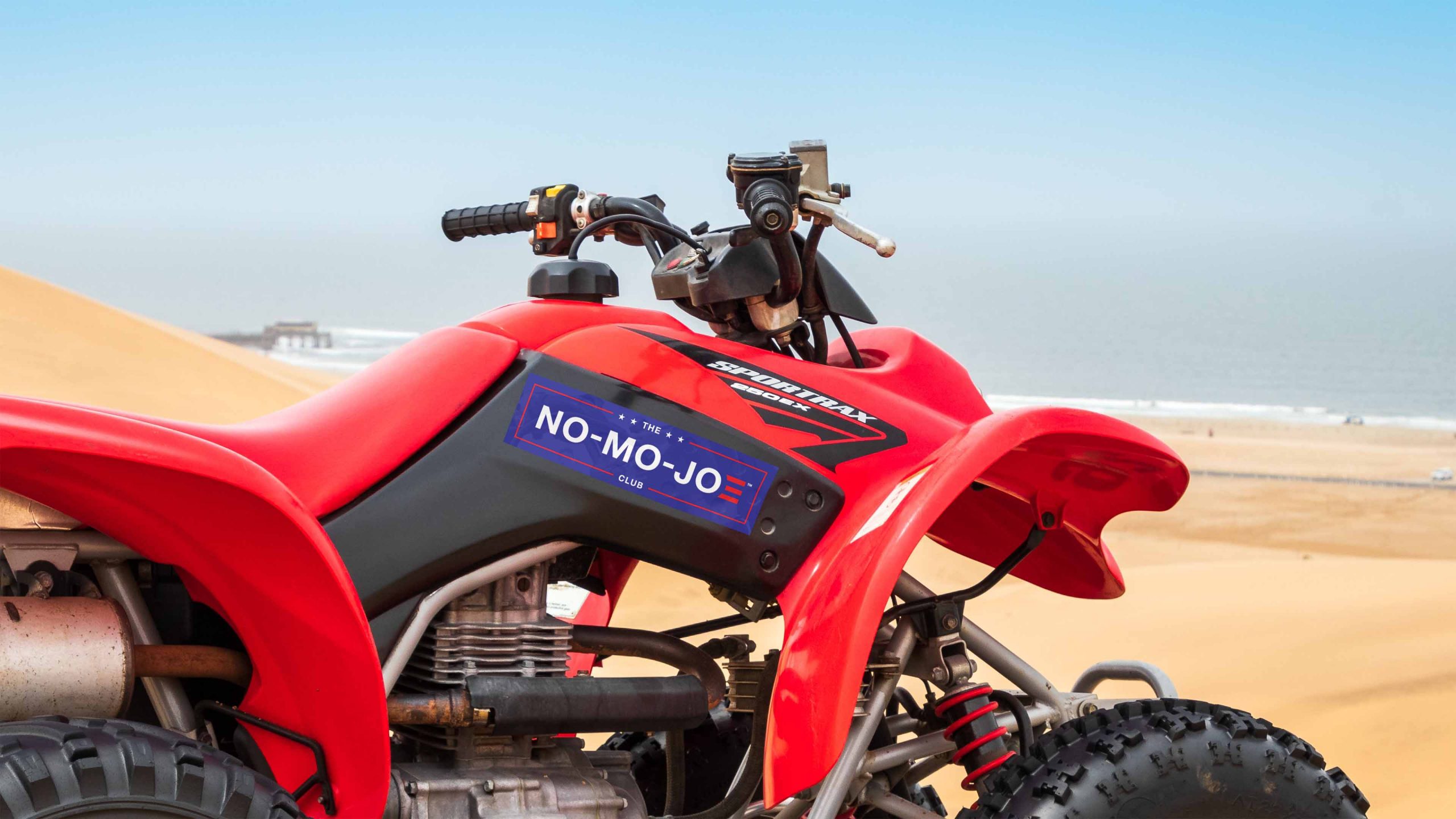 The No-Mo-Joe Club™ sticker on side panel of red four wheeler in dunes near the beach.