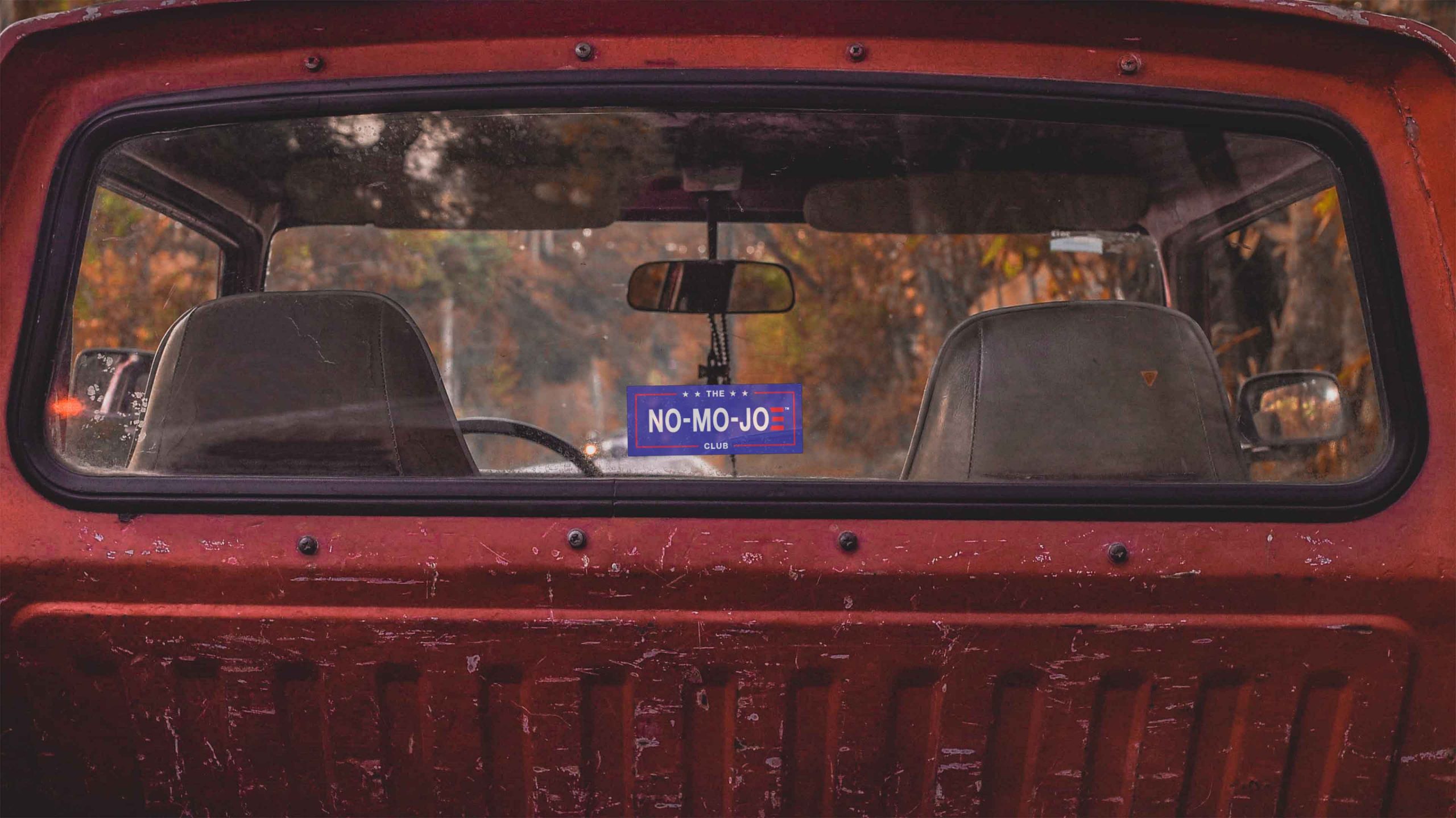The No-Mo-Joe Club™ window cling on the back window of a scratched, dark red pickup with a rosary hanging from the rear view mirror