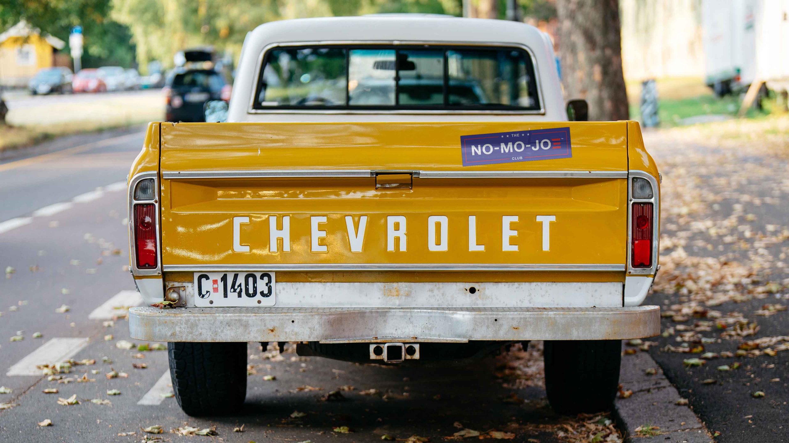 Vintage, Yellow Chevrolet pickup truck with The No-Mo-Joe Club™ bumper sticker parked on the side of the road on a fall day