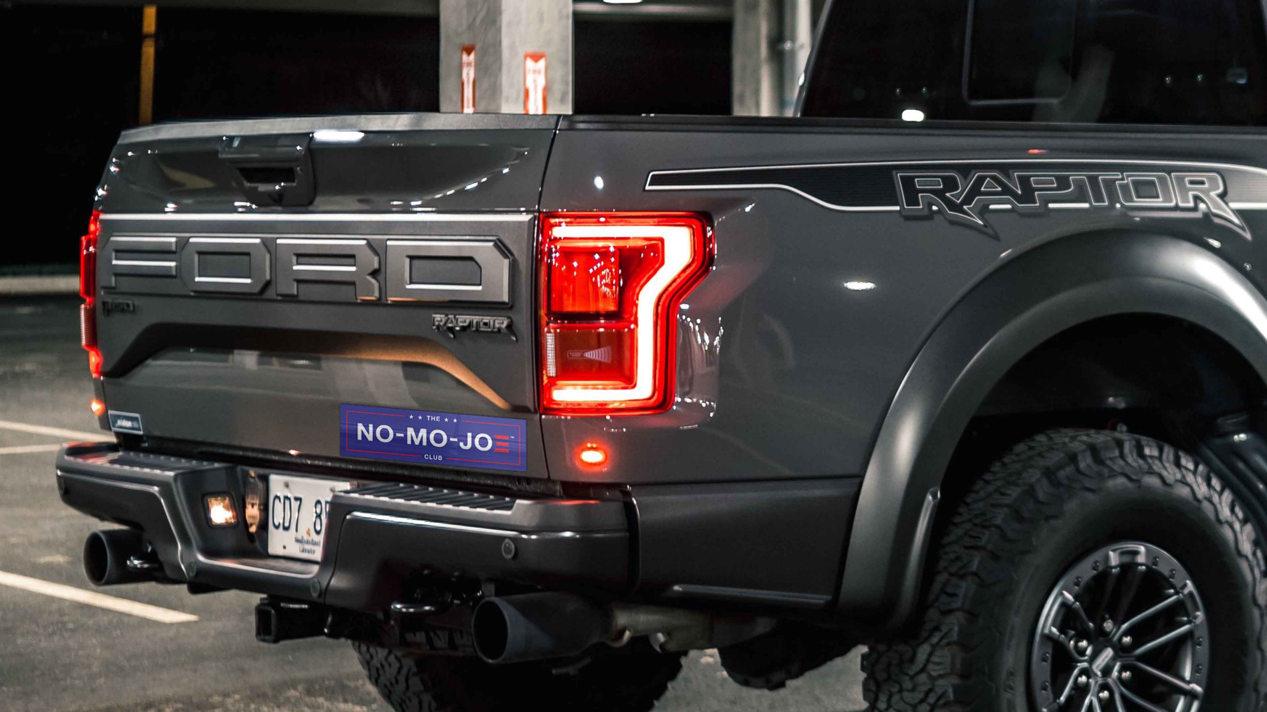 Tail gate of a Gray, loaded Ford Raptor with a The No-Mo-Joe Club™ bumper sticker in a parking garage.