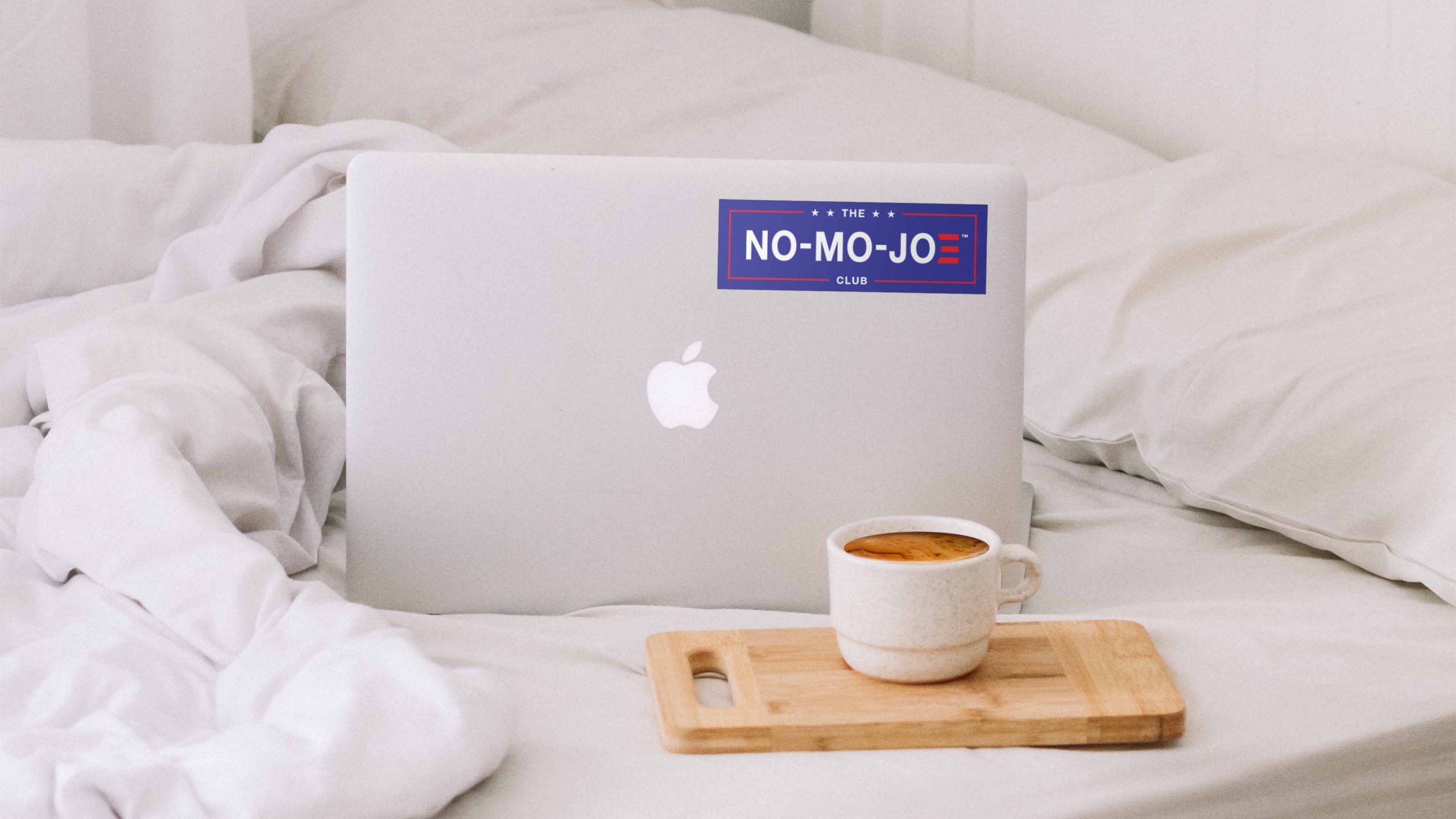 The No-Mo-Joe Club™ sticker on laptop on bed with white sheets, pillows, duvet and small wooden board with cup of fresh brewed coffee