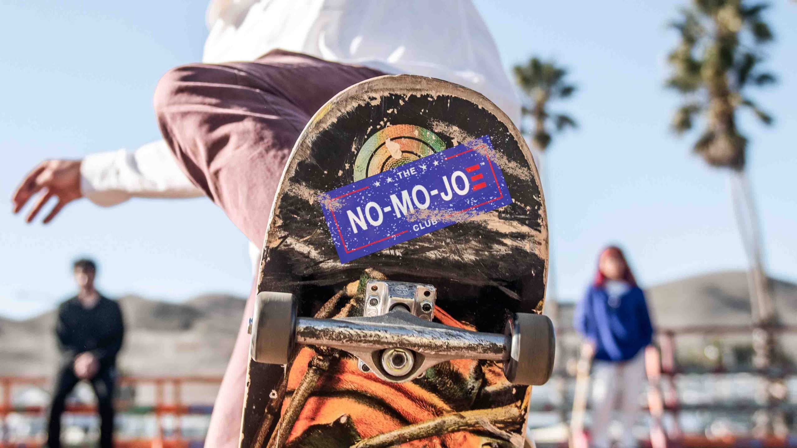 The No-Mo-Joe Club™ sticker on the bottom of skateboard during trick with palm trees in background.